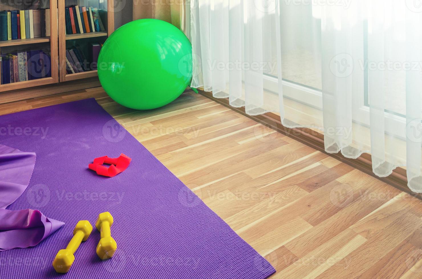 Sports equipment on floor in living room, violet yoga mat, yellow dumbbells, red rubber resistance band and green rubber fitness aerobic ball photo