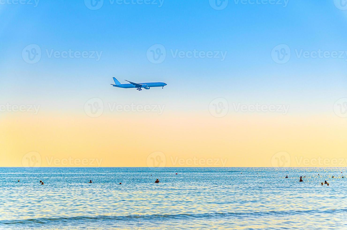 Airplane flying low above sea and people tourists swimming in water, clear blue orange sky at sunset, plane preparing to land photo