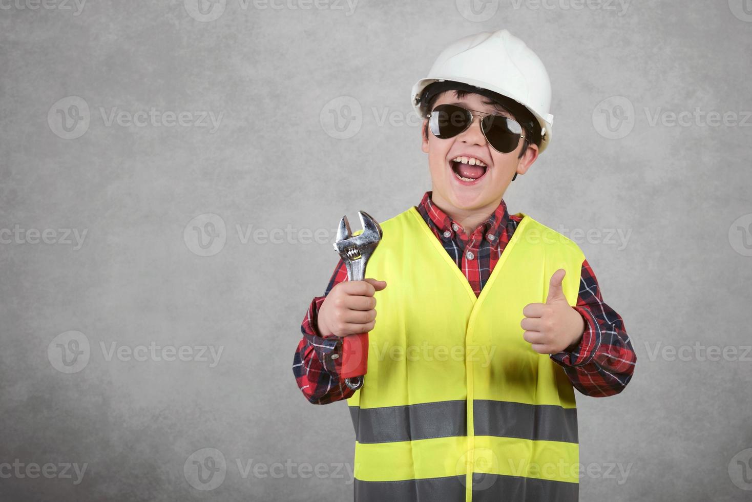 niño pequeño trabajador de la construcción con casco blanco y gafas de sol y sosteniendo una llave foto