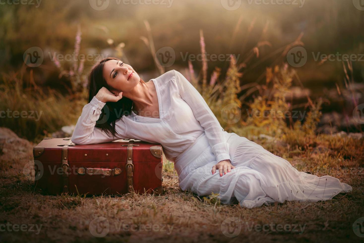 young woman sitting in the field with suitcase photo