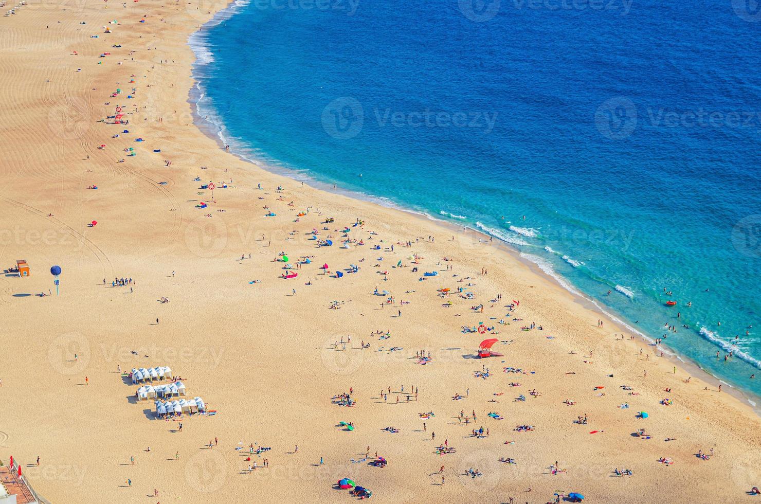 vista aérea superior de la playa de arena con gente turistas tomando el sol y agua turquesa azul del océano atlántico, praia da nazare foto