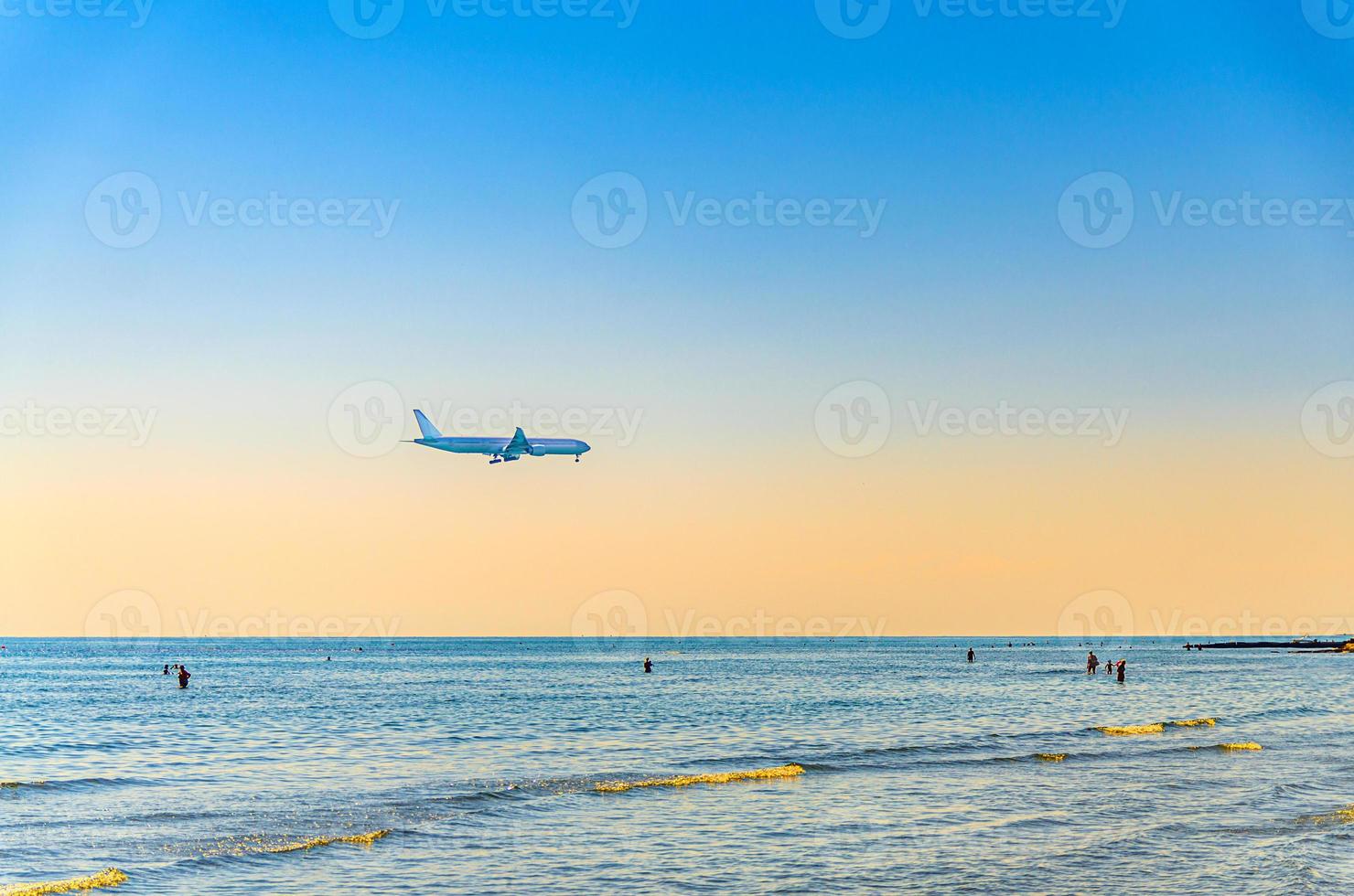 Airplane flying low above sea and people tourists swimming in water, clear blue orange sky at sunset, plane preparing to land photo