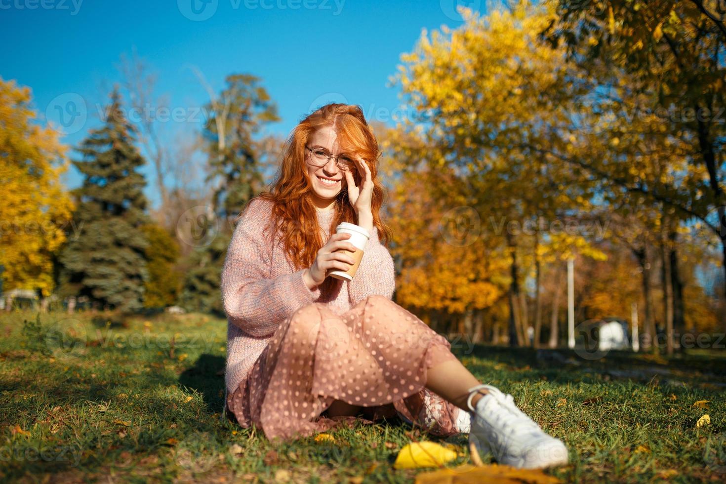 retratos de una encantadora chica pelirroja con gafas y una cara bonita. chica posando en el parque de otoño en un suéter y una falda de color coral. foto