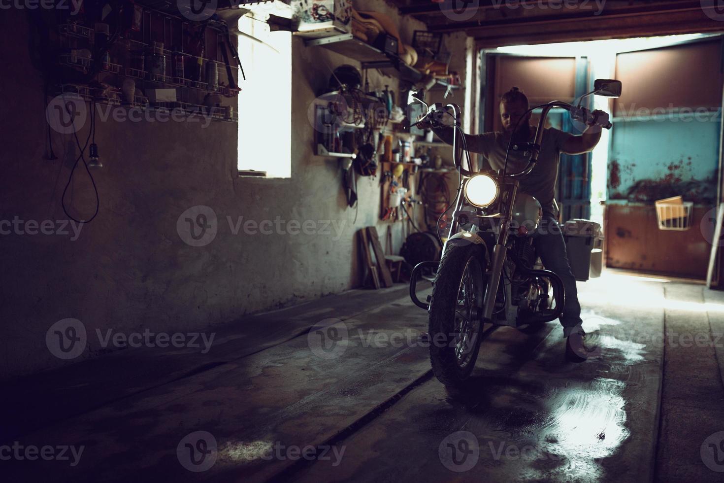 un hombre brutal y guapo con barba sentado en una motocicleta en su garaje foto
