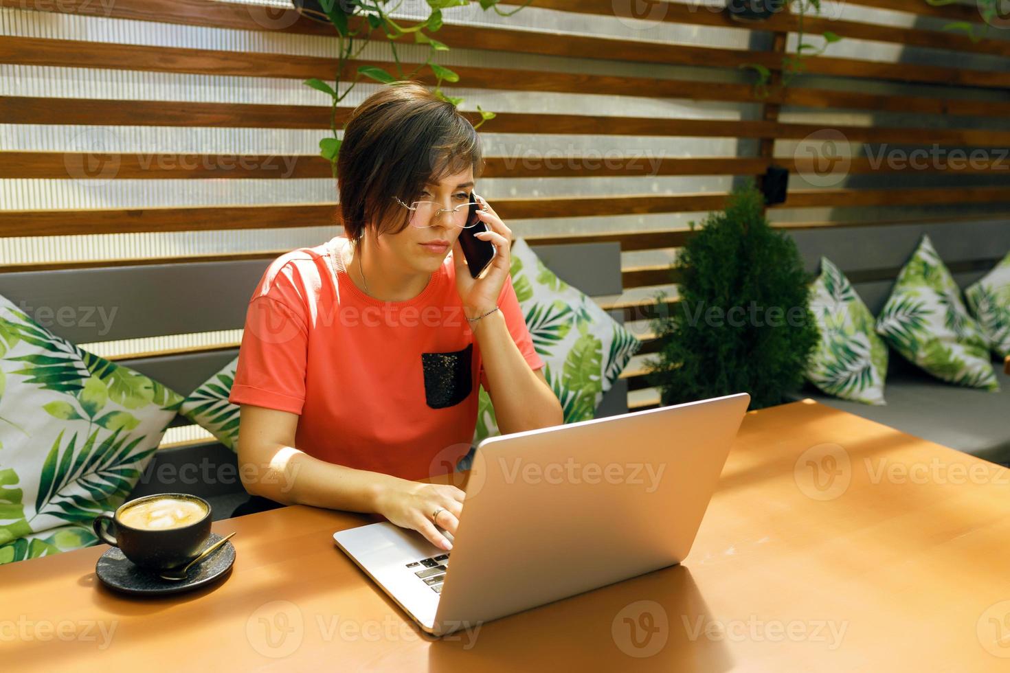Portrait of confident mature professional woman in glasses, a coral T-shirt sitting on summer terrace in cafe, using laptop computer for work, laughing happily indoors photo