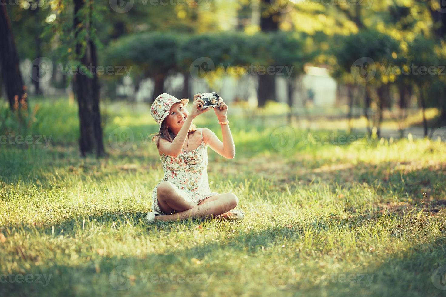 little girl is played by photo camera sitting on grass in park. Doing Selfie and photographing the world around