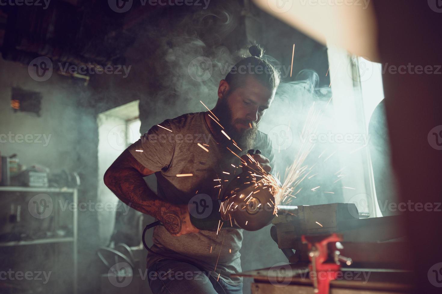 Handsome brutal male with a beard repairing a motorcycle in his garage working with a circular saw. In the garage a lot of sparks and smoke from sawing photo