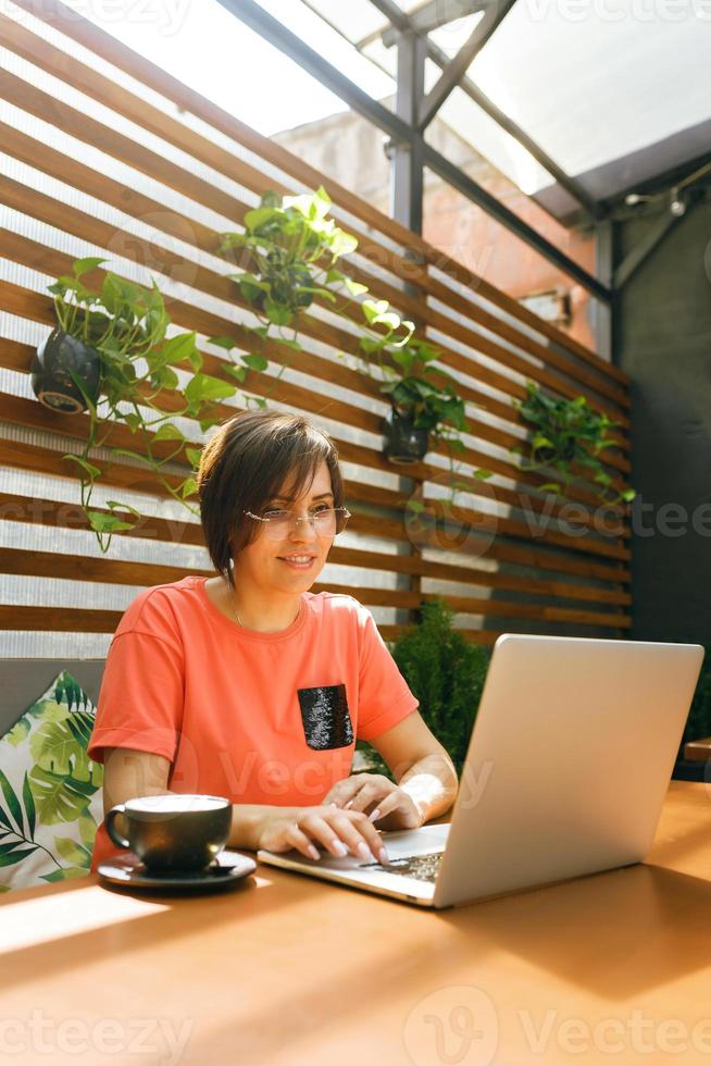 Portrait of confident mature professional woman in glasses, a coral T-shirt sitting on summer terrace in cafe, using laptop computer for work, laughing happily indoors photo