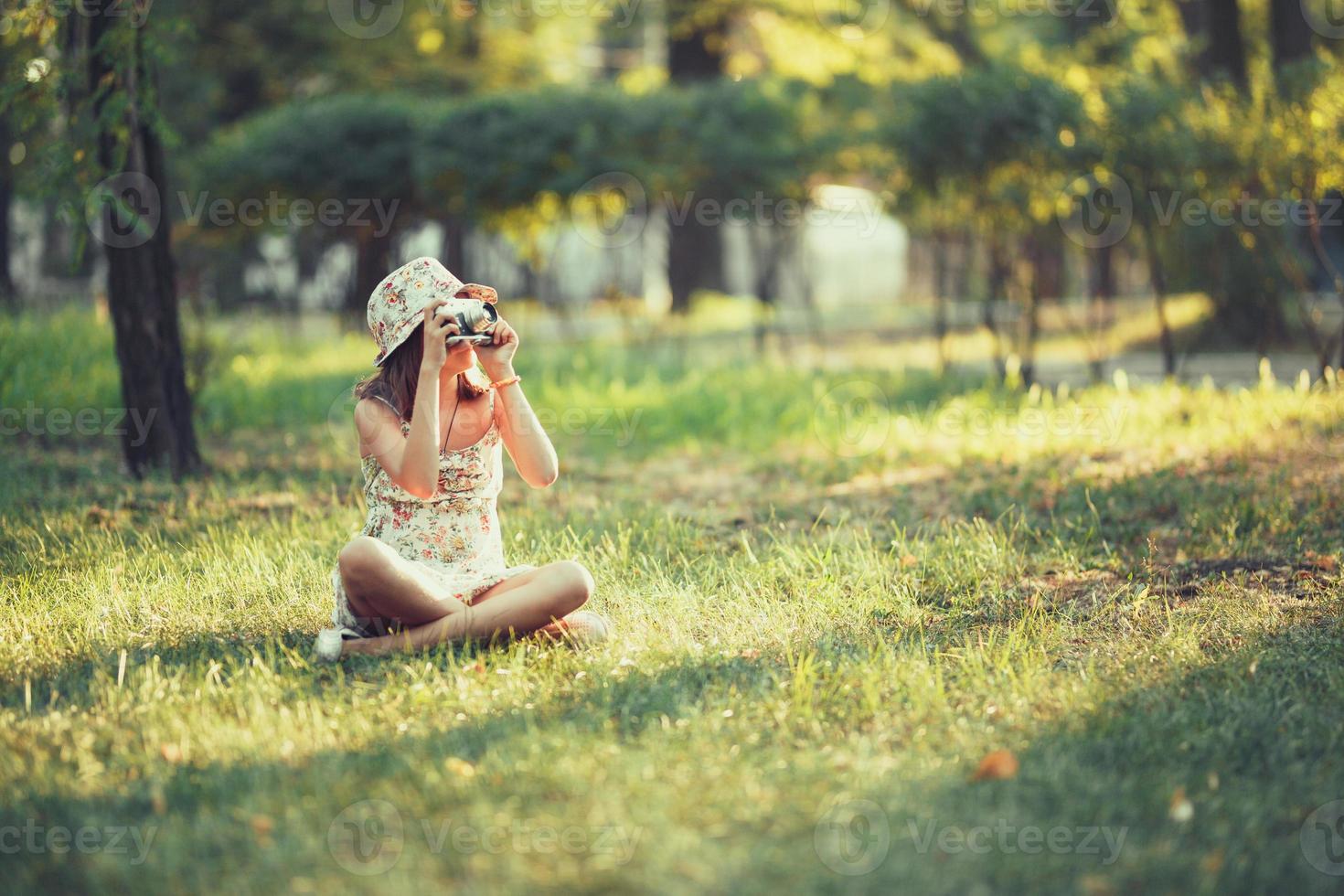 la niña pequeña es interpretada por una cámara fotográfica sentada en el césped en el parque. haciendo selfie y fotografiando el mundo alrededor foto