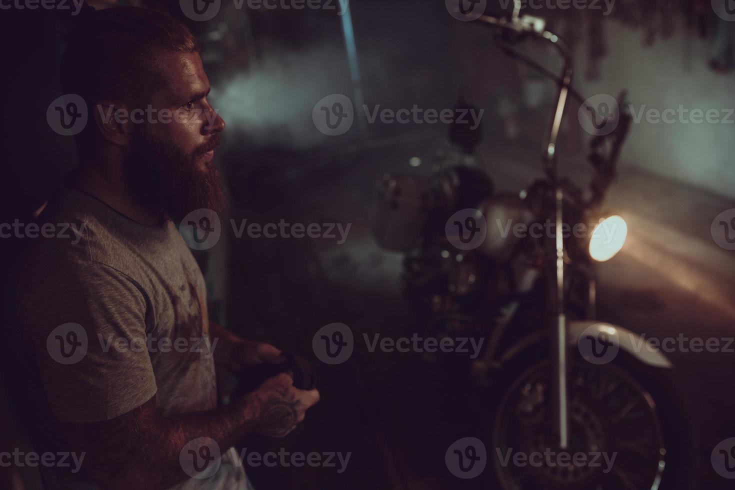 Handsome brutal man with a beard is standing in his garage against the background of a motorcycle and looking away photo