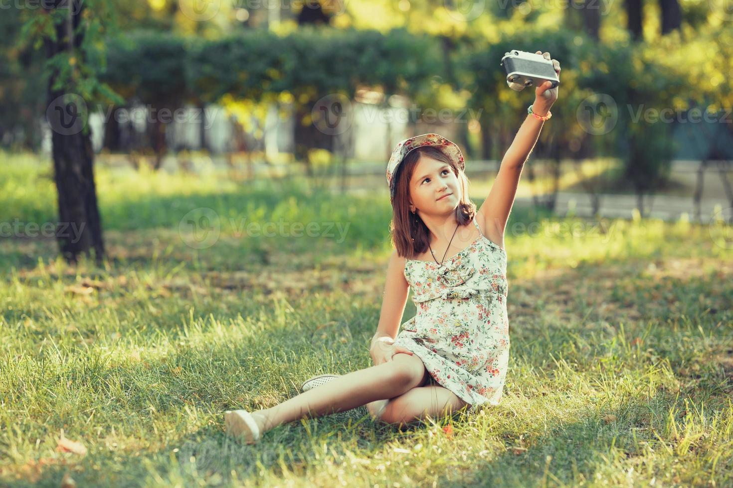little girl is played by photo camera sitting on grass in park. Doing Selfie and photographing the world around