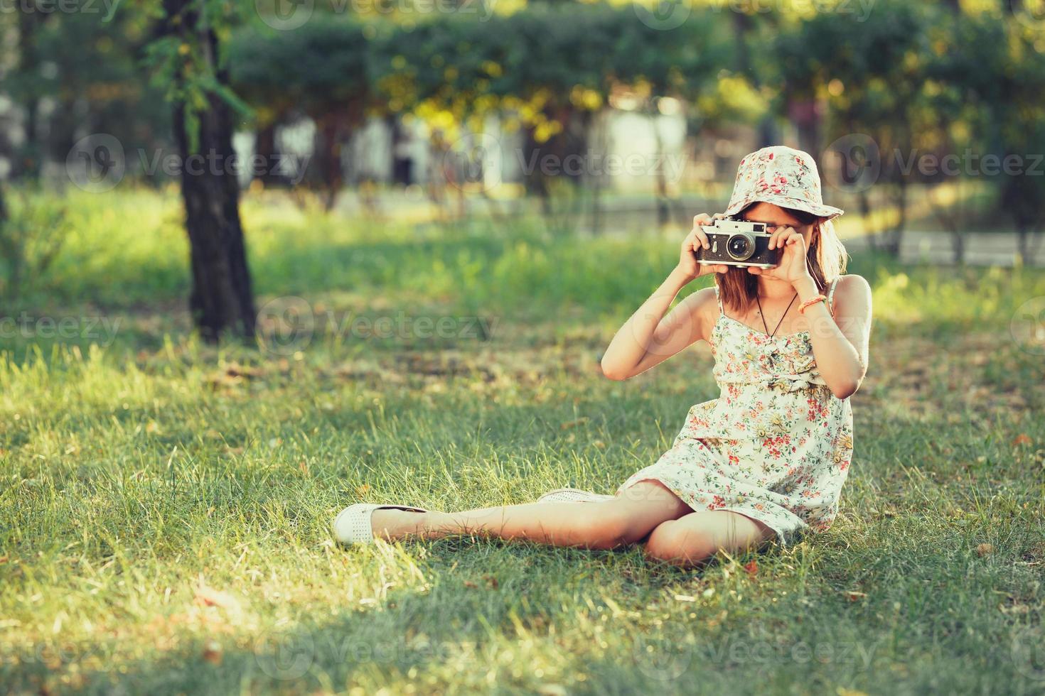 la niña pequeña es interpretada por una cámara fotográfica sentada en el césped en el parque. haciendo selfie y fotografiando el mundo alrededor foto