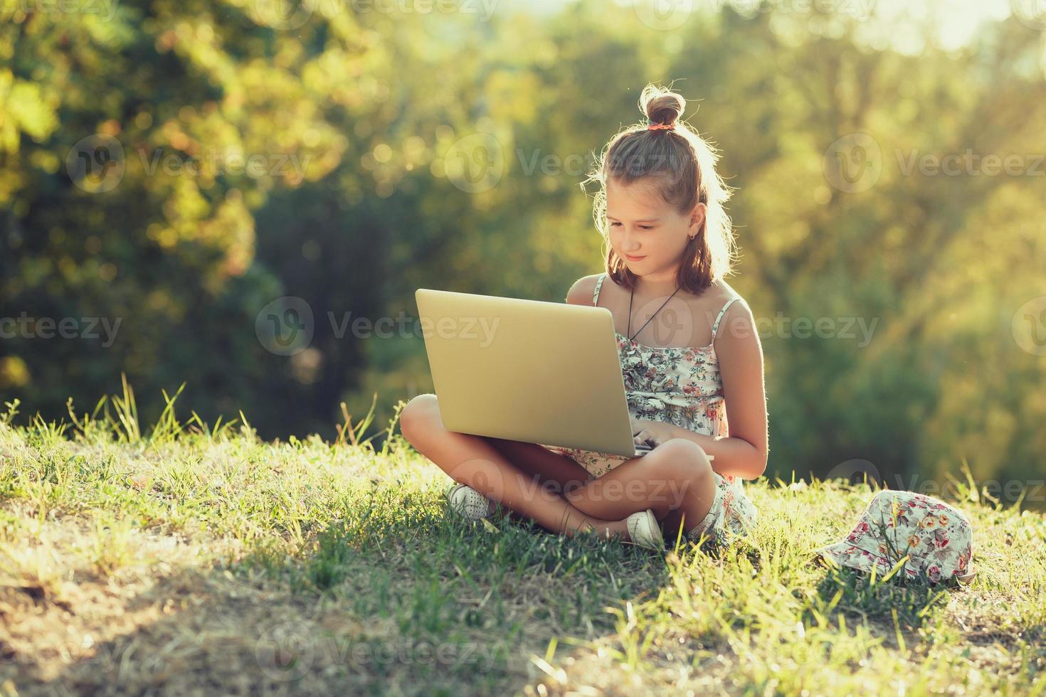 la niña está hablando en una computadora portátil mientras se sienta en el césped bajo el sol. vestido con un sarafan y un sombrero foto