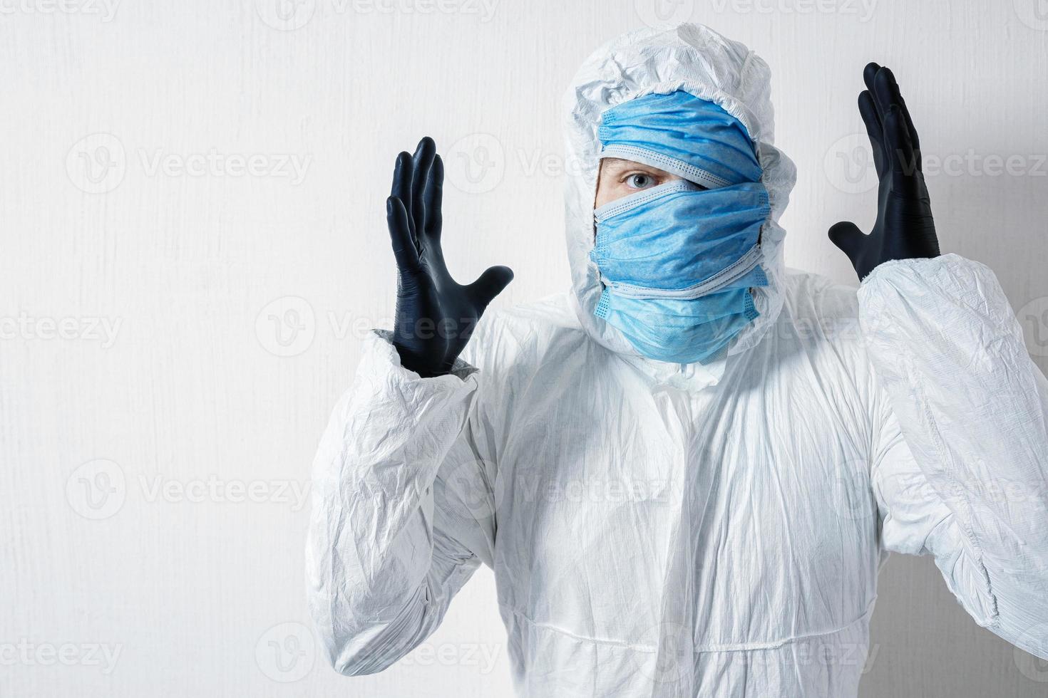 a frightened man in a protective suit hung with medical masks against a white wall. The horrors of the epidemic, the danger of the coronavirus, scientist depicts surprise and despair photo