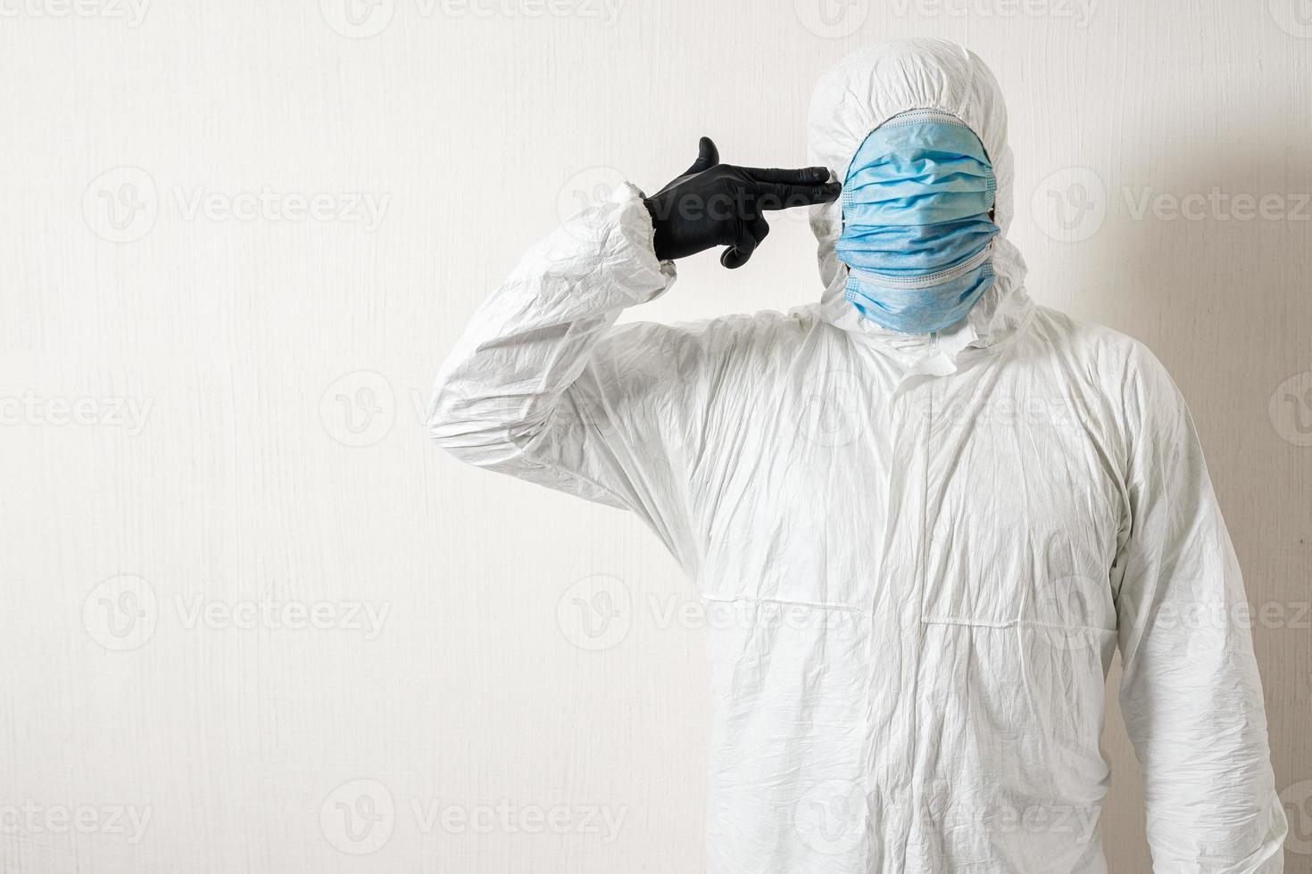 un hombre con un traje protector colgado con máscaras médicas posando contra un fondo de pared mostrando varios gestos con los dedos el científico le apunta con una pistola a la sien foto