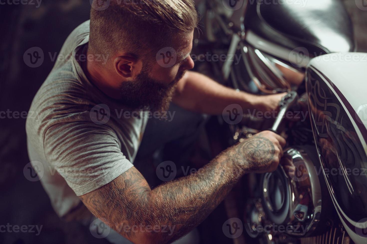 un apuesto hombre barbudo con chaqueta de cuero y gafas de sol está sentado en la motocicleta en el taller de reparación foto