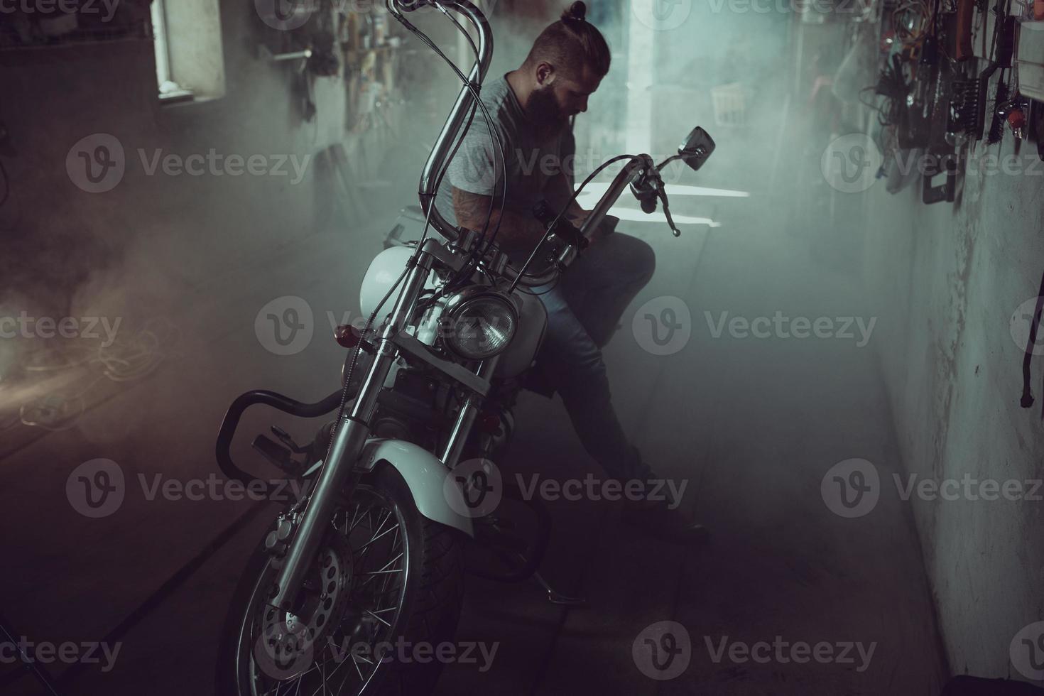 un hombre guapo y brutal con barba sentado en una motocicleta en su garaje, limpiándose las manos y mirando hacia un lado foto