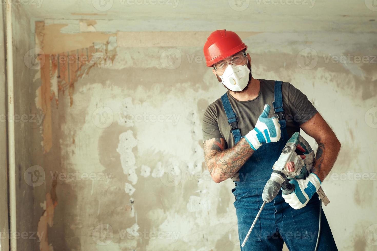 a builder with a perforator poses against the background of a wall with empty space from text The builder is dressed in a protective suit and helmet photo