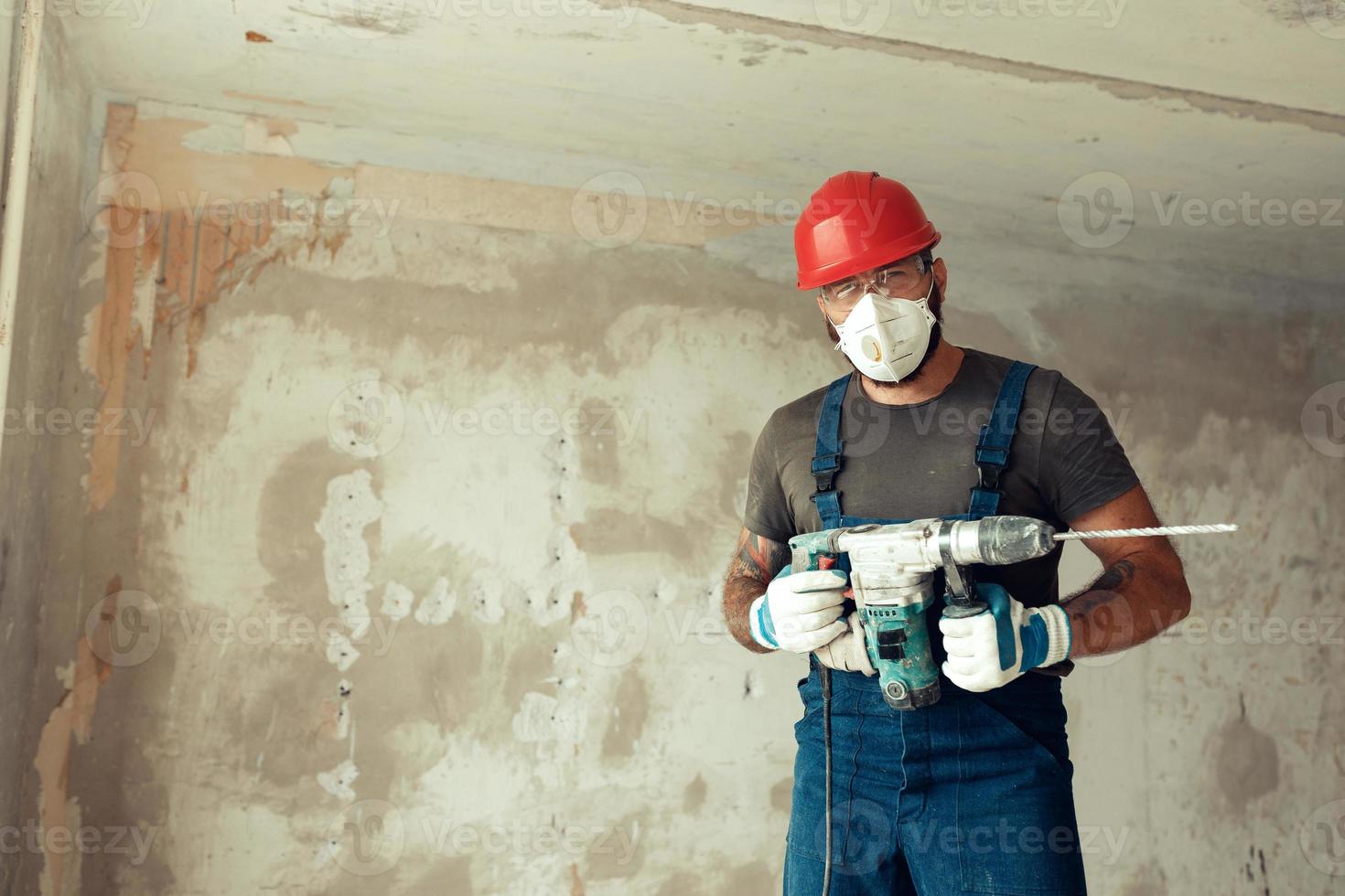 a builder with a perforator poses against the background of a wall with empty space from text The builder is dressed in a protective suit and helmet photo