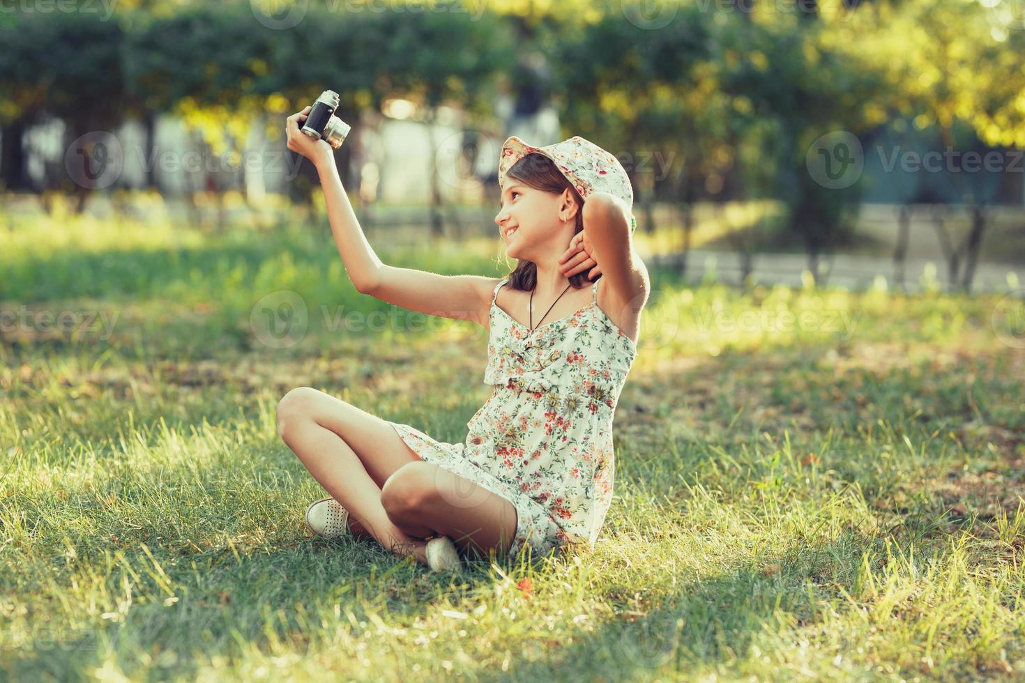 la niña pequeña es interpretada por una cámara fotográfica sentada en el césped en el parque. haciendo selfie y fotografiando el mundo alrededor foto