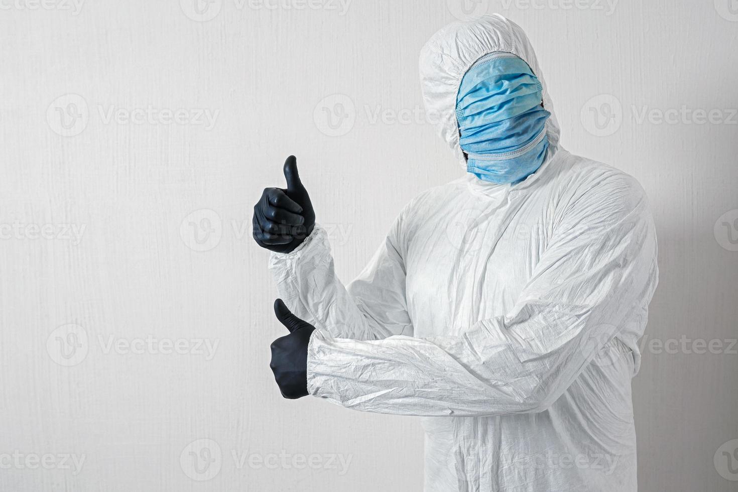 a man in a protective suit hung with medical masks posing against a wall background showing various gestures with his fingers, the scientist shows a thumb up on both hands photo