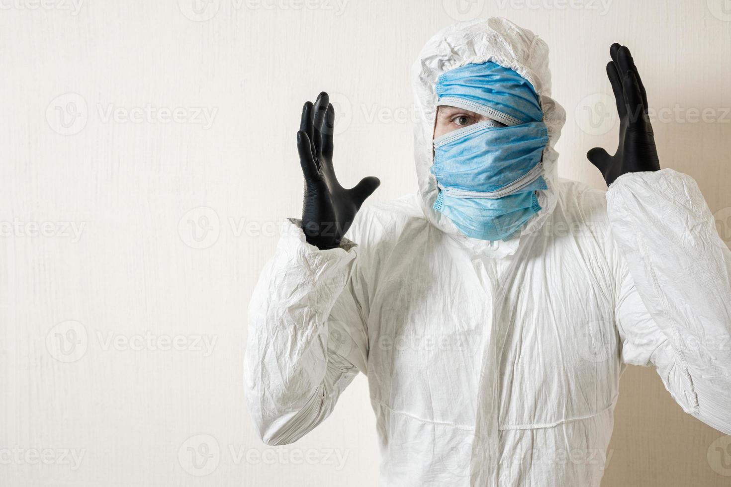 a frightened man in a protective suit hung with medical masks against a white wall. The horrors of the epidemic, the danger of the coronavirus, scientist depicts surprise and despair photo
