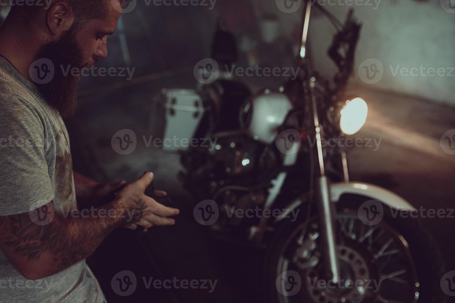 Handsome brutal man with a beard is standing in his garage against the background of a motorcycle and looking away photo