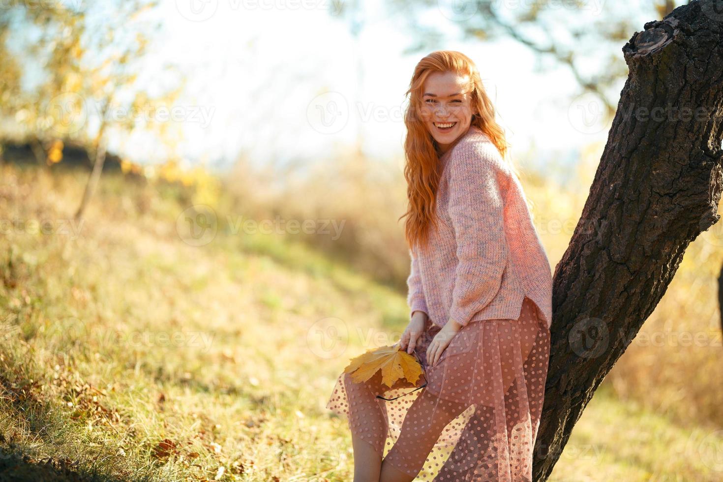 Portraits of a charming red-haired girl with a cute face. Girl posing in autumn park in a sweater and a coral-colored skirt. In the hands of a girl a yellow leaf photo