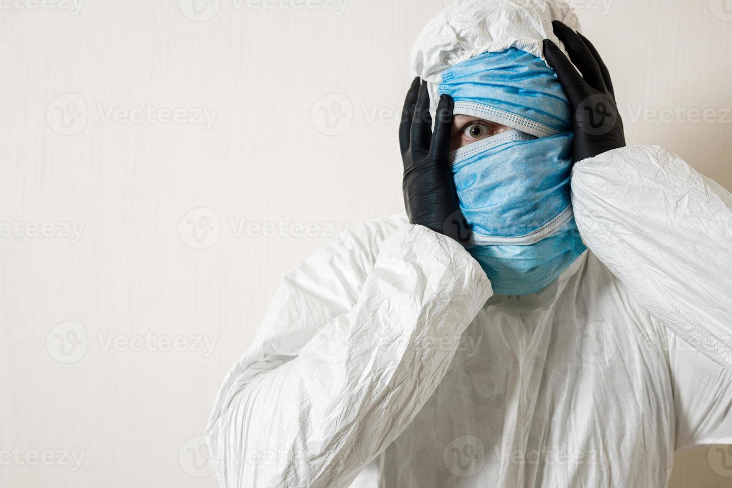 a frightened man in a protective suit hung with medical masks depicts horror against a white wall. The horrors of the epidemic, the danger of the coronavirus photo