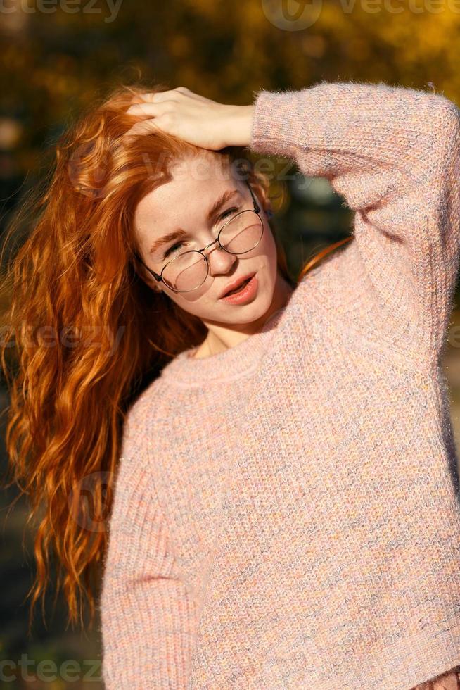 retratos de una encantadora chica pelirroja con gafas y cara linda. chica posando en el parque de otoño con un suéter y una falda de color coral. la niña tiene un estado de ánimo maravilloso foto