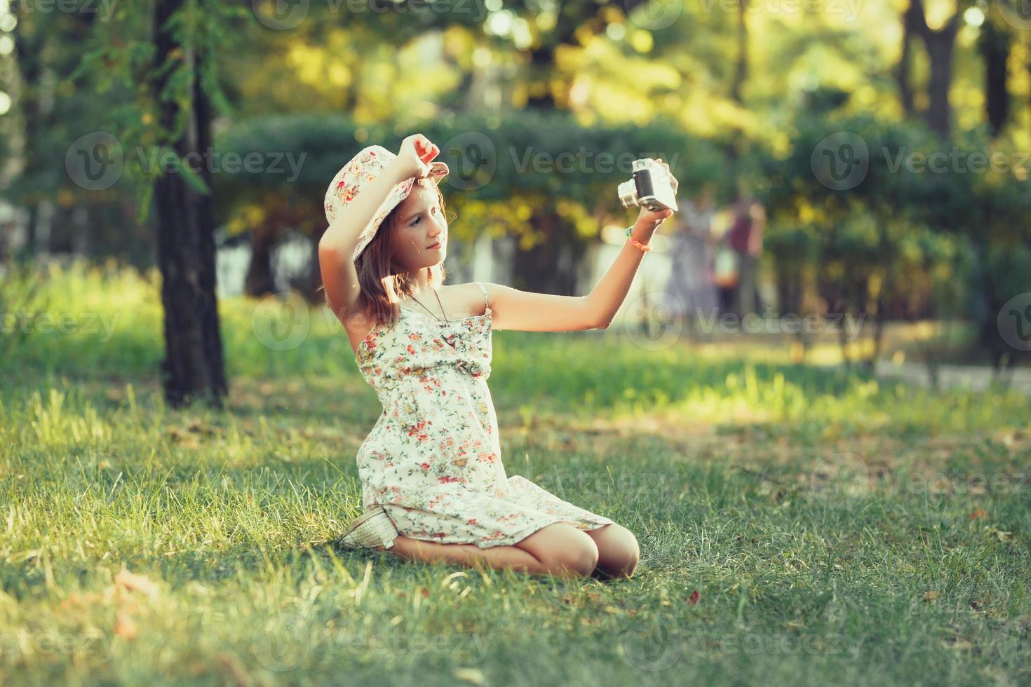 little girl is played by photo camera sitting on grass in park. Doing Selfie and photographing the world around