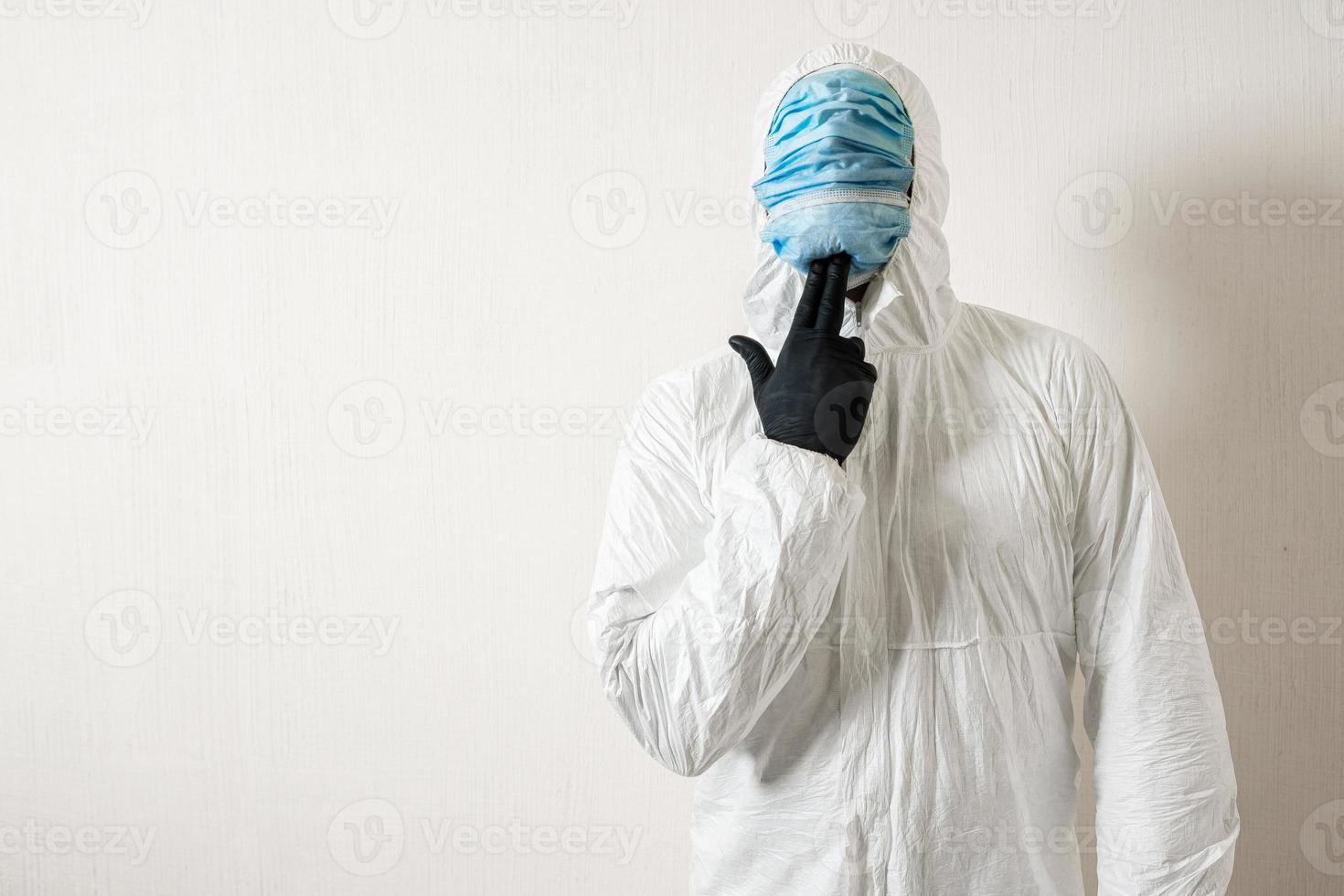 a man in a protective suit hung with medical masks posing against a wall background showing various gestures with his fingers photo