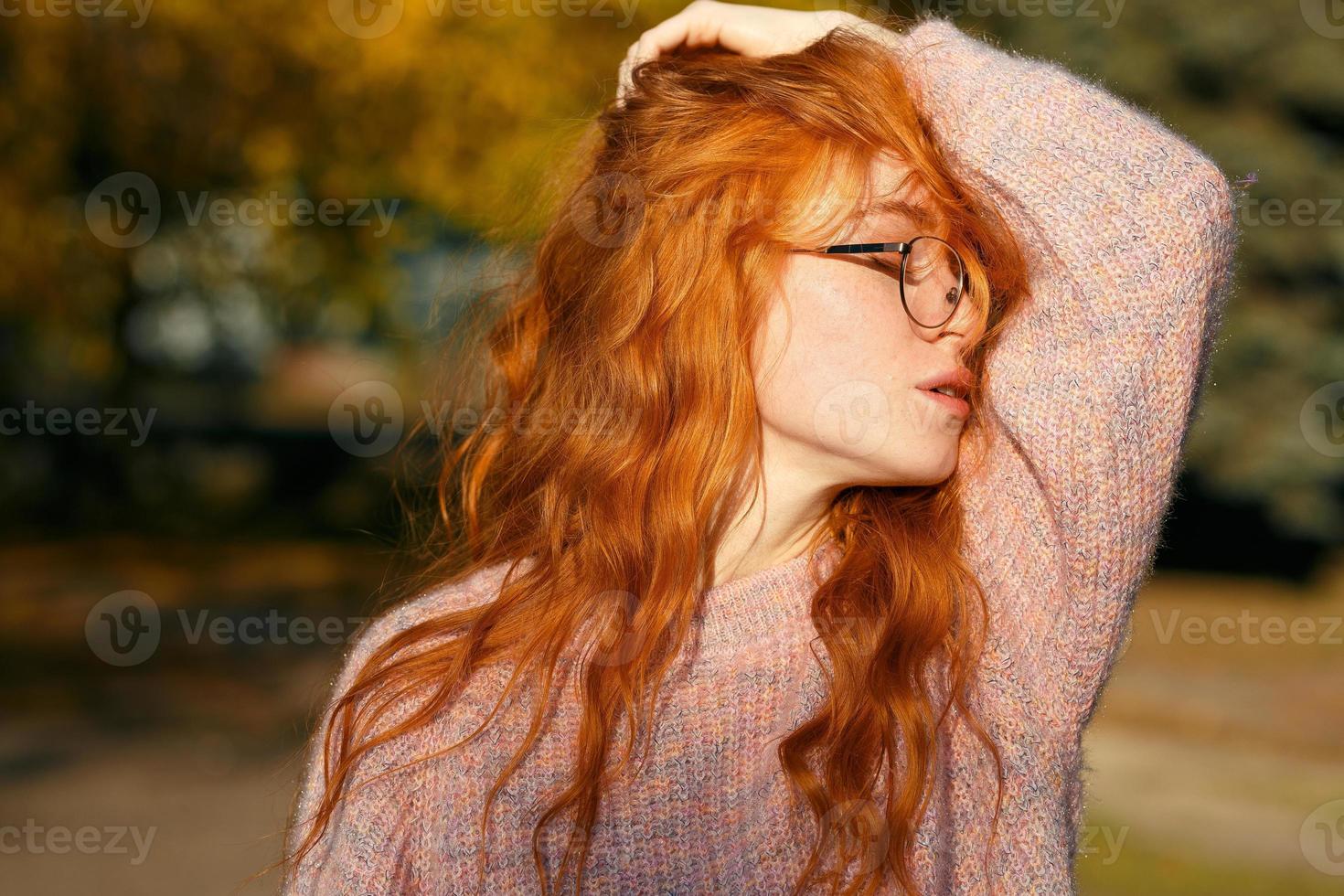 retratos de una encantadora chica pelirroja con gafas y cara linda. chica posando en el parque de otoño con un suéter y una falda de color coral. la niña tiene un estado de ánimo maravilloso foto