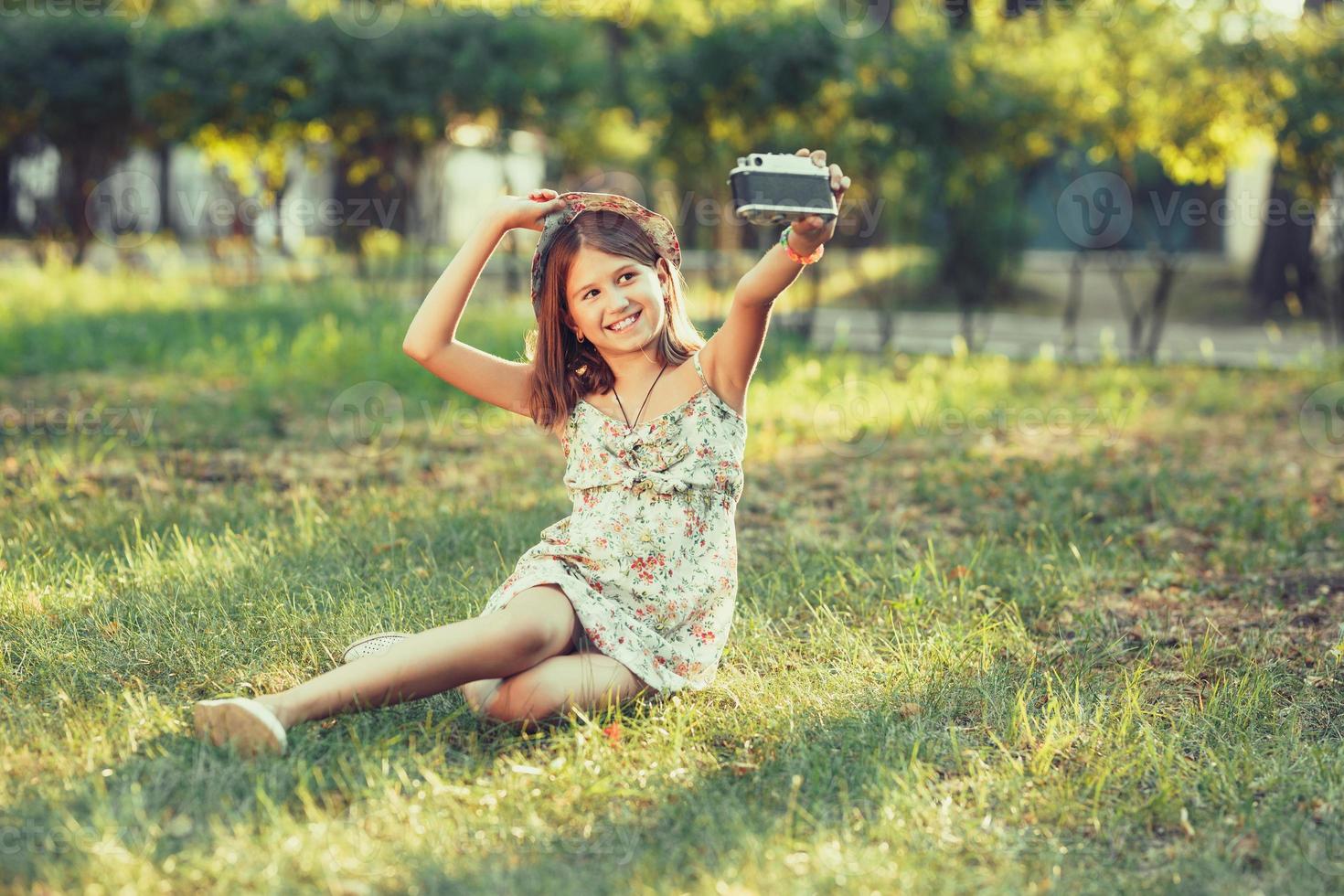 little girl is played by photo camera sitting on grass in park. Doing Selfie and photographing the world around