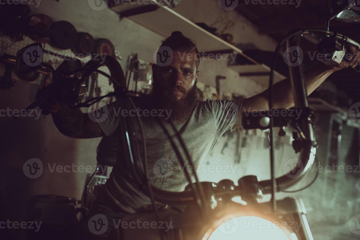 Handsome brutal man with a beard sitting on a motorcycle in his garage, wiping his hands and looking to the side photo
