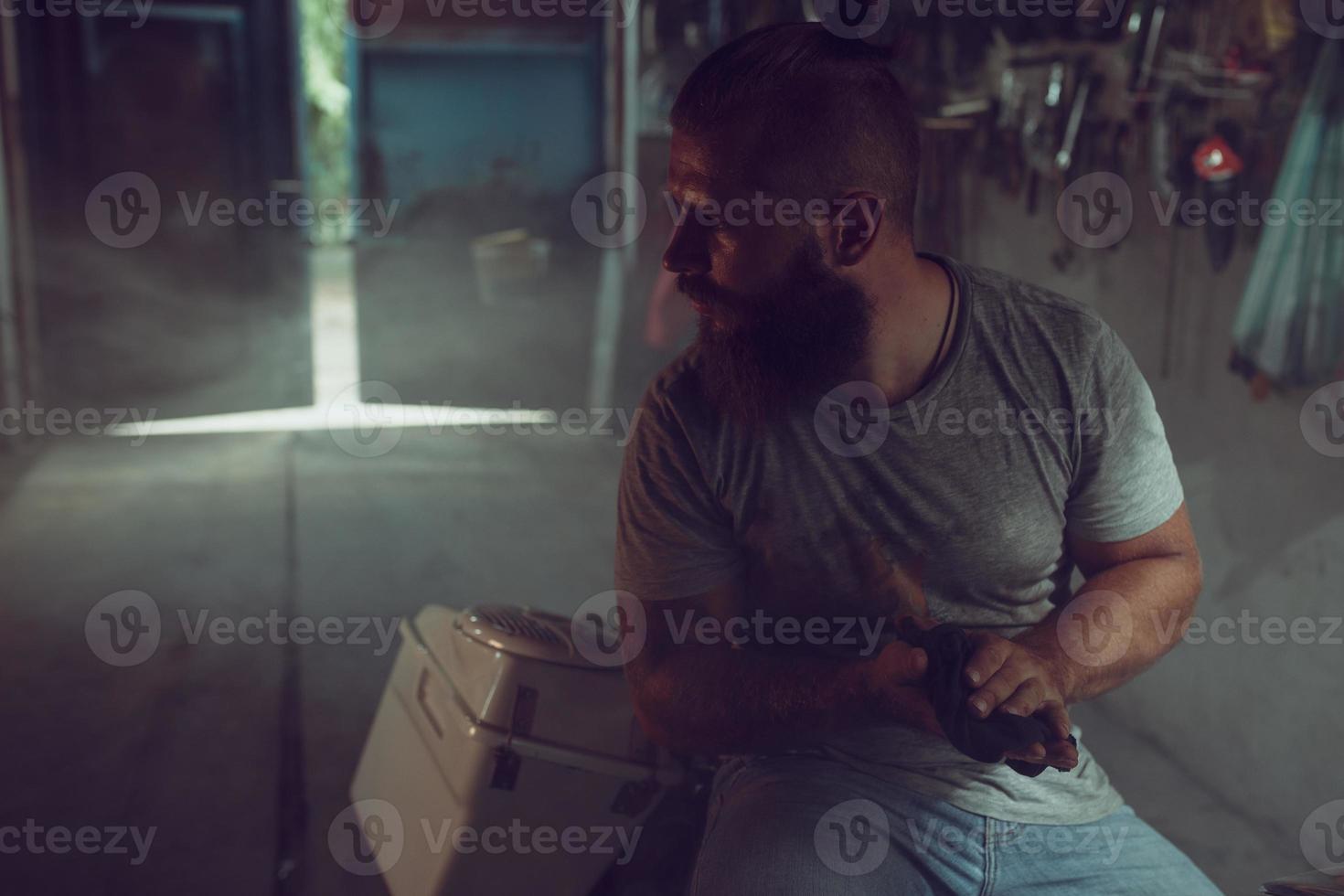 Handsome brutal man with a beard sitting on a motorcycle in his garage, wiping his hands and looking to the side photo