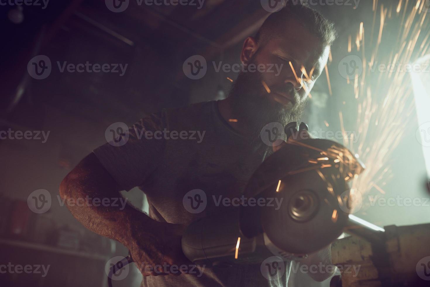 Handsome brutal male with a beard repairing a motorcycle in his garage working with a circular saw. In the garage a lot of sparks and smoke from sawing photo