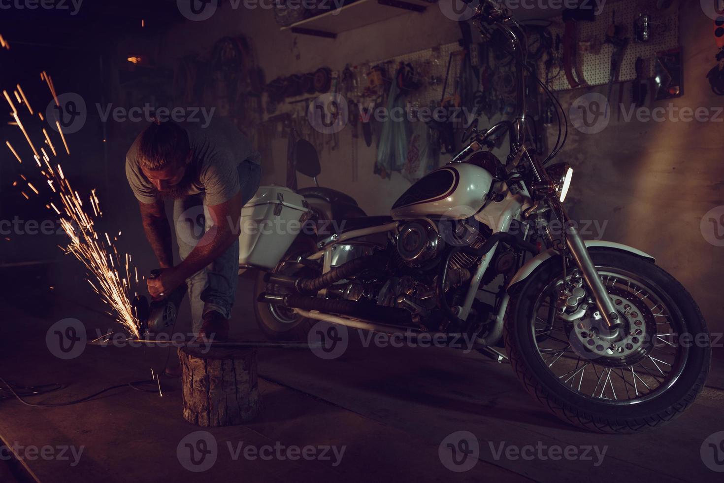 Handsome brutal male with a beard repairing a motorcycle in his garage working with a circular saw. In the garage a lot of sparks and smoke from sawing photo