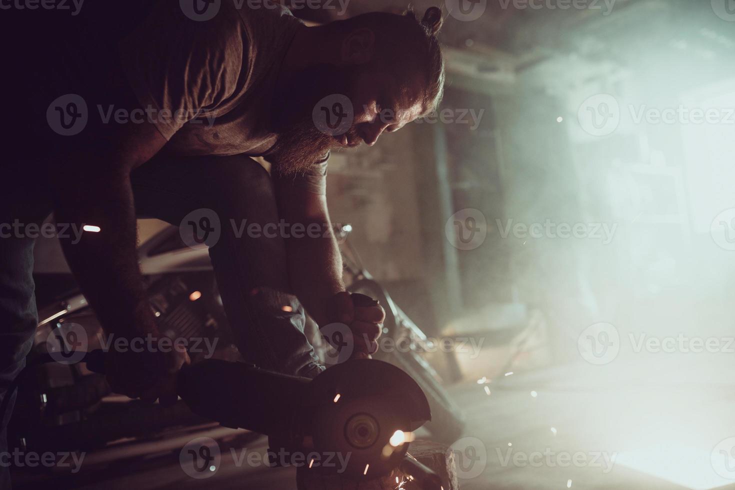 Handsome brutal male with a beard repairing a motorcycle in his garage working with a circular saw. In the garage a lot of sparks and smoke from sawing photo