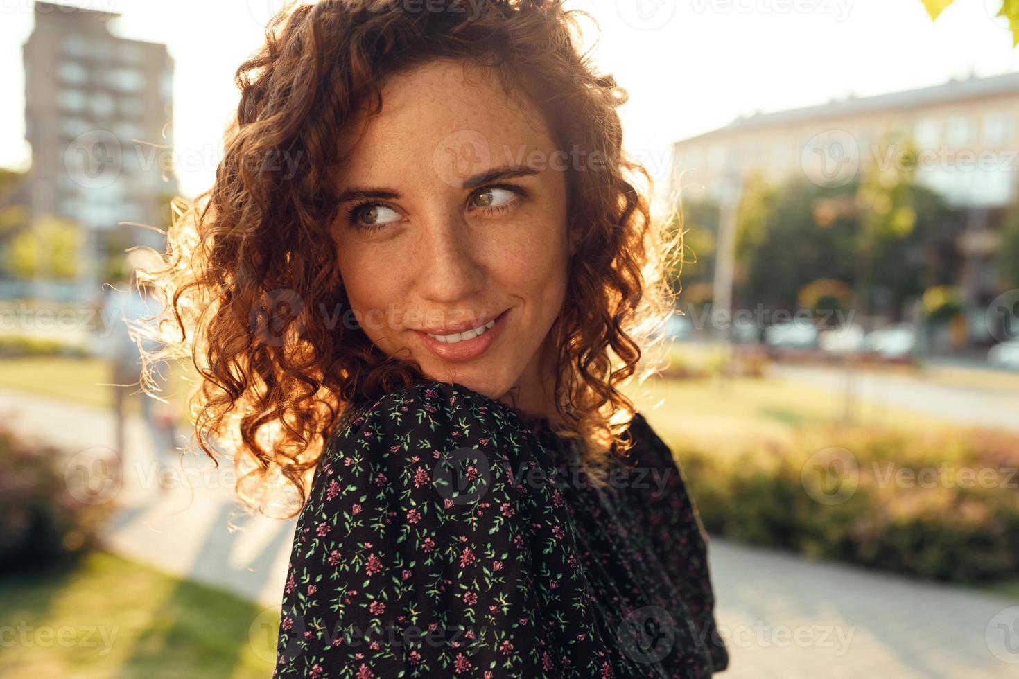 charming curly red-haired girl with freckles in dress poses for the camera in the city center showing different facial emotions photo