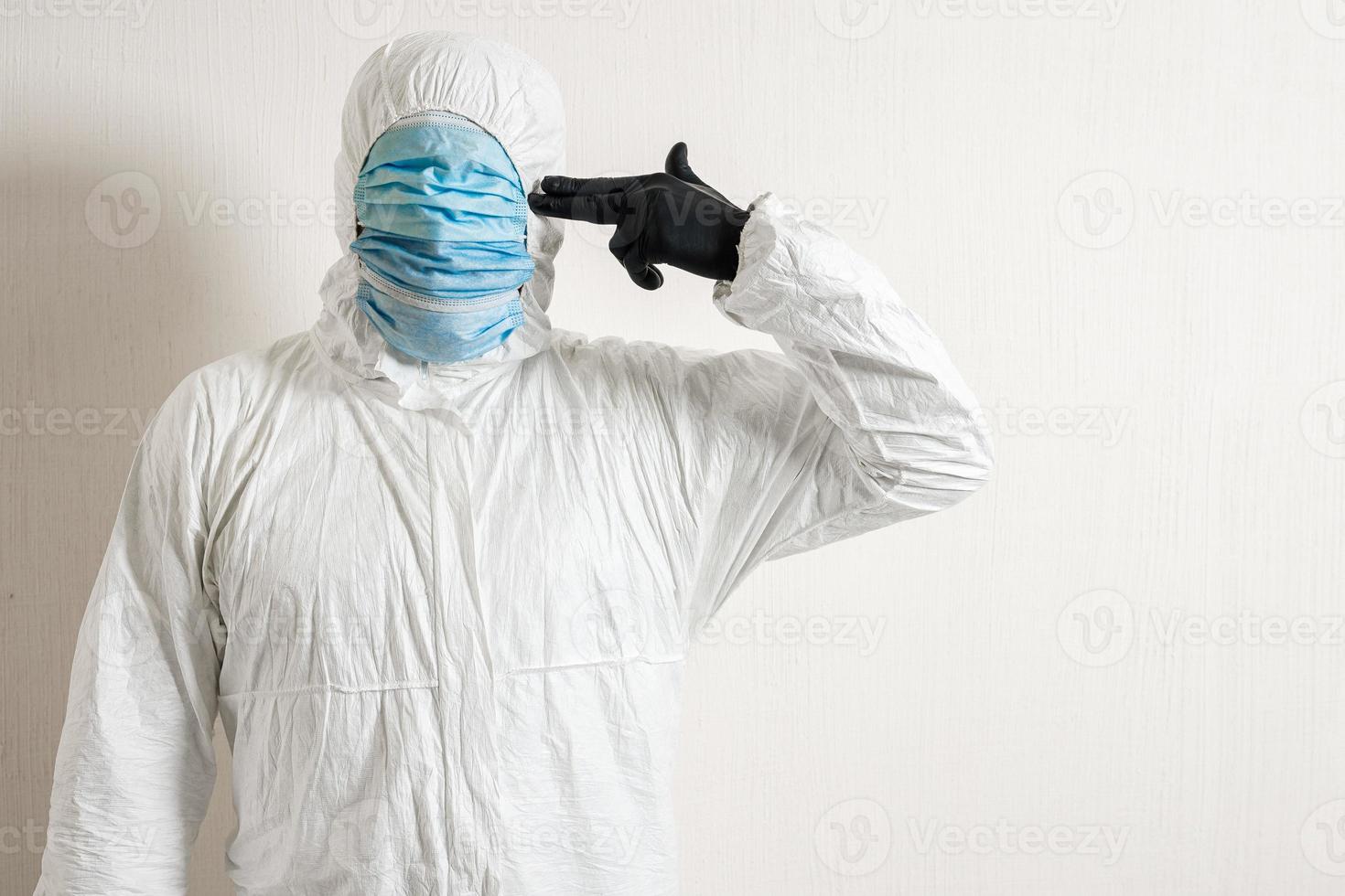 a man in a protective suit hung with medical masks posing against a wall background showing various gestures with his fingers the scientist gestures a pistol to his temple photo