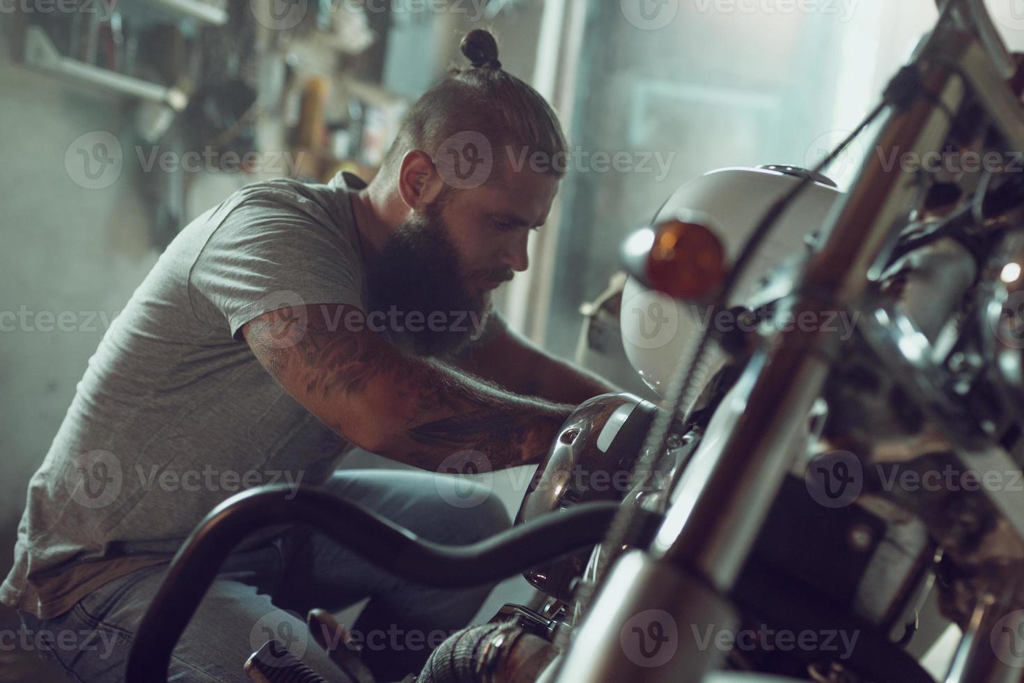 hombre guapo con barba reparando su motocicleta en el garaje. un hombre vestido con jeans y una camiseta foto