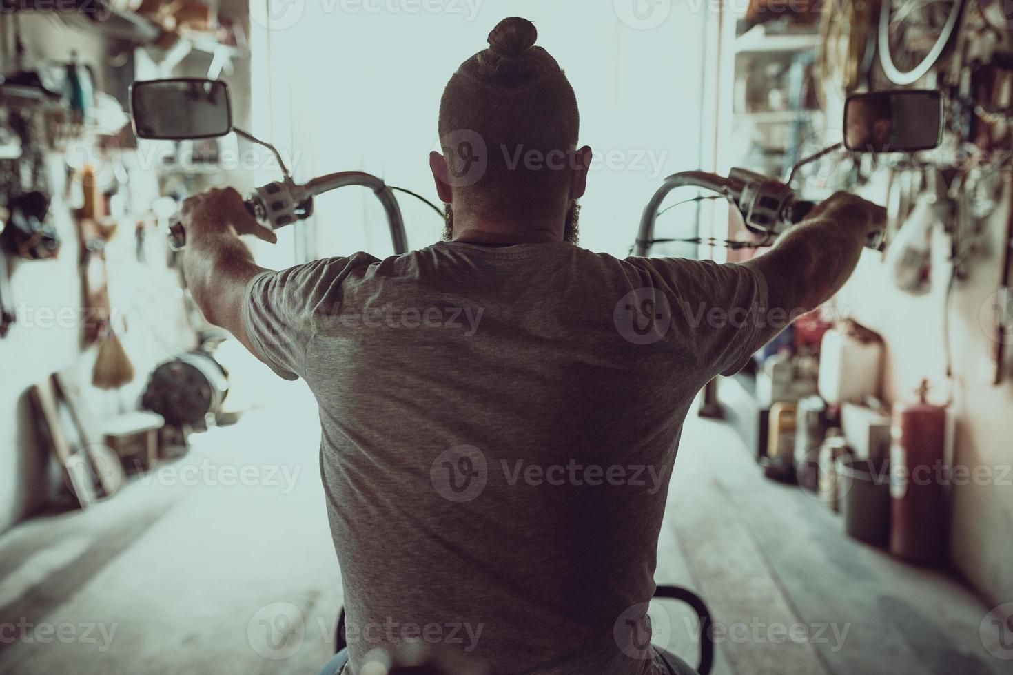Handsome brutal man with a beard sitting on a motorcycle in his garage, view from the back photo