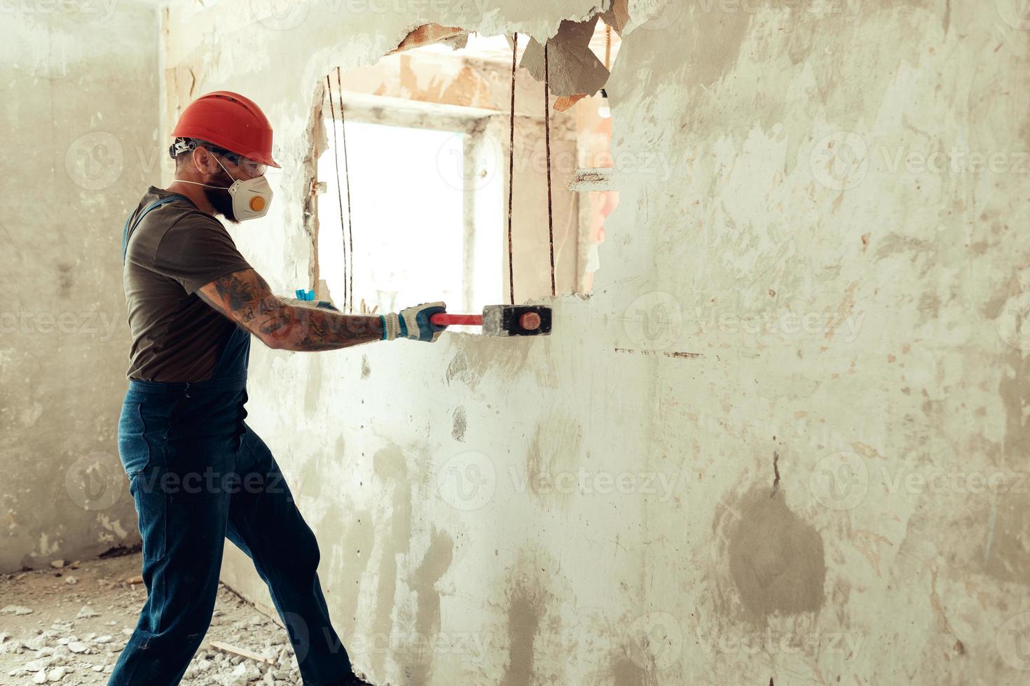 el constructor con un martillo en las manos rompe la pared de cemento el constructor está vestido con un traje protector y un casco foto