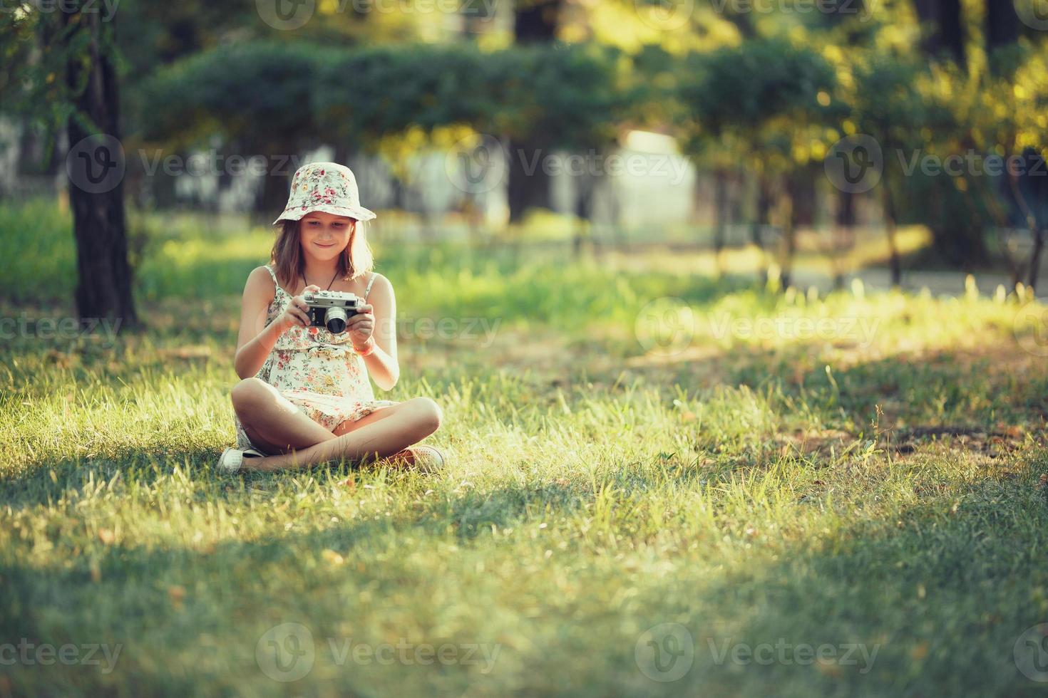 little girl is played by photo camera sitting on grass in park. Doing Selfie and photographing the world around