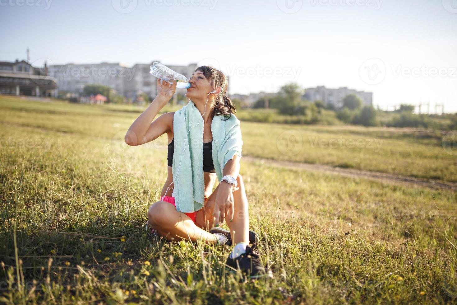 chica deportiva bebiendo agua después del deporte. niña sentada en la hierba. edificio en el fondo foto