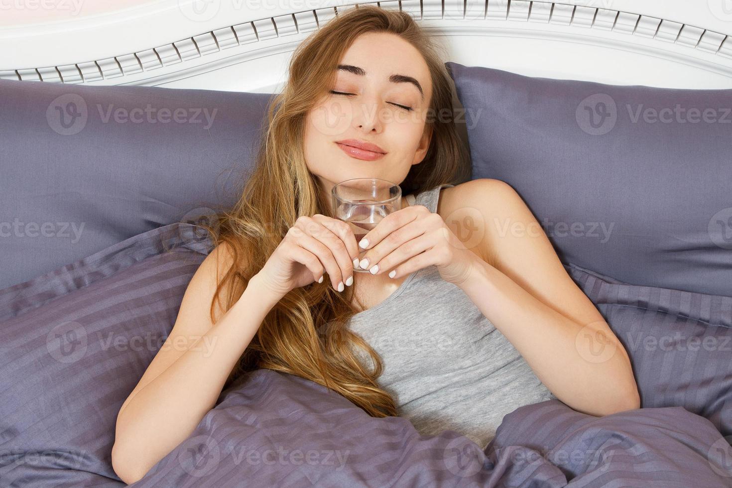 Happy young woman holding a glass of water and lying in bed in the bedroom - female hangover photo