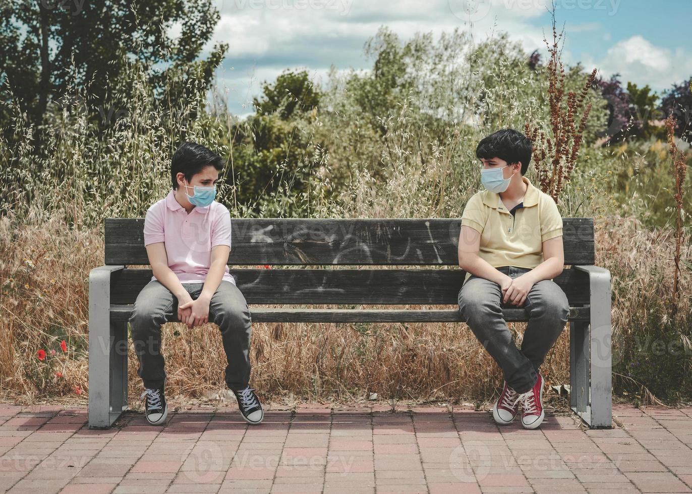 dos niños con mascarilla médica mantienen el distanciamiento social para evitar la propagación del virus sentados en un banco foto
