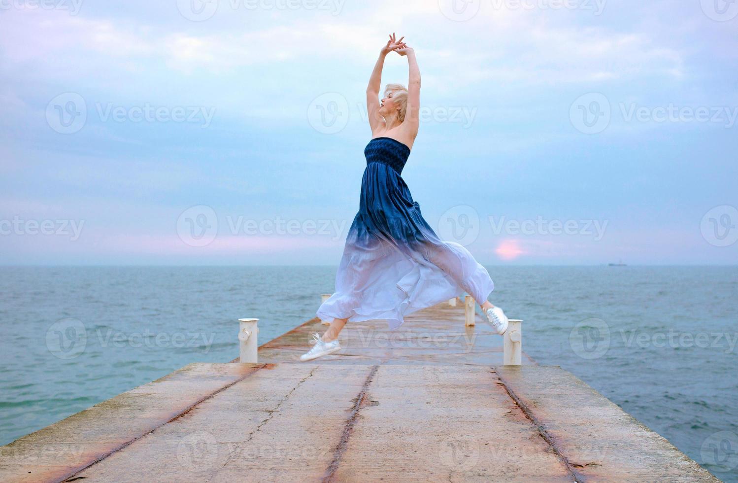 mujer rubia blanca caucásica con pelo corto en vestido largo y zapatillas felices de estar embarazada y saltando junto al mar al amanecer foto