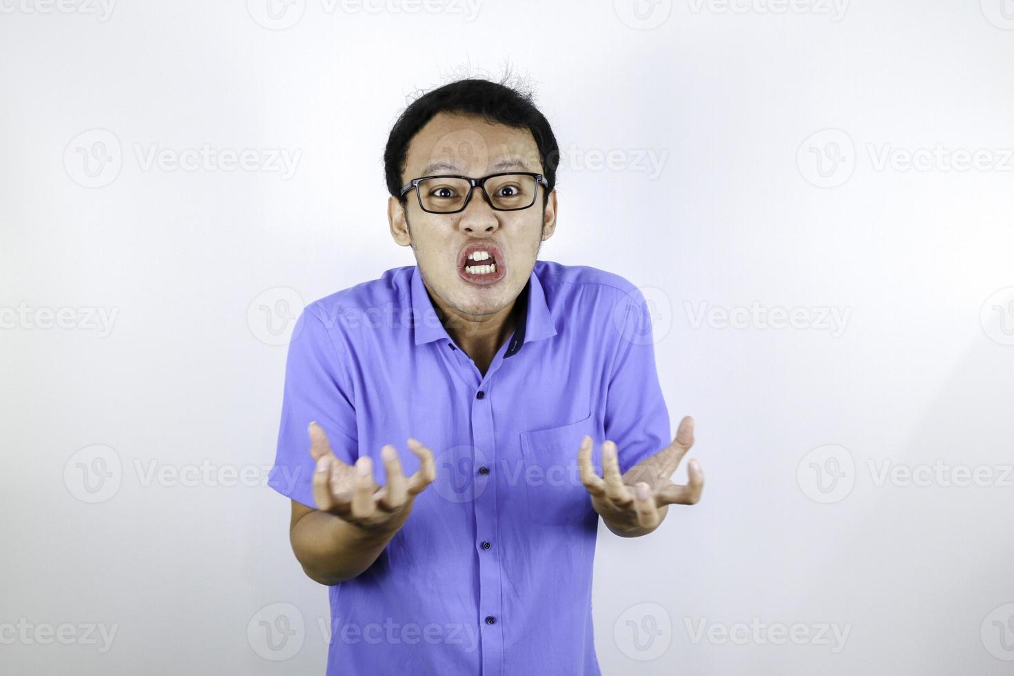 El joven asiático que usa camisa azul es una cara graciosa y enojada con gritos y señalando con el dedo a la cámara aislada sobre el fondo blanco. foto